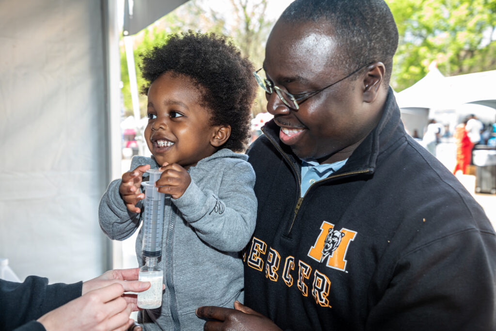 Child holding a tube with liquid, smiling while being held by an adult. Both are under a tent at an outdoor event.