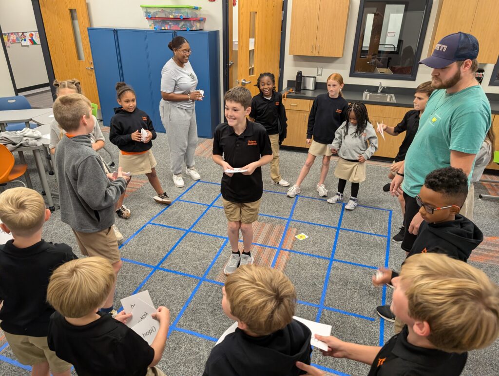 Children and adults engage in a grid-based activity in a classroom, holding cards and interacting with each other.