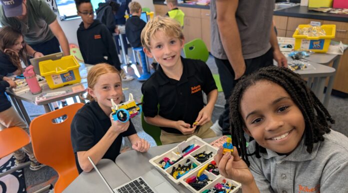 Mercerians teach coding skills to Roberts Academy students Three children smile while assembling building blocks and using a laptop in a classroom setting, with others in the background.