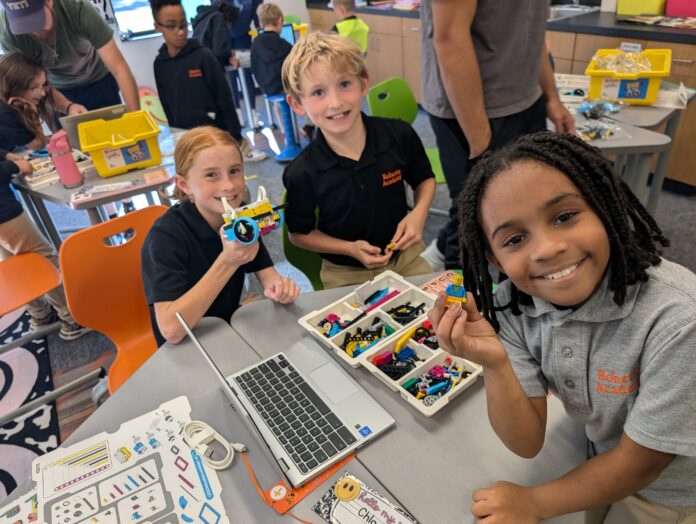 tech quest 1 Three children smile while assembling building blocks and using a laptop in a classroom setting, with others in the background.