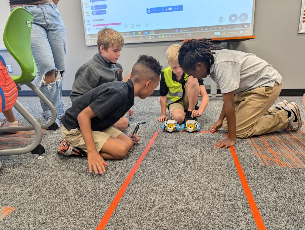 Four children kneeling on a carpeted floor, playing with small robotic cars on a track in a classroom setting.