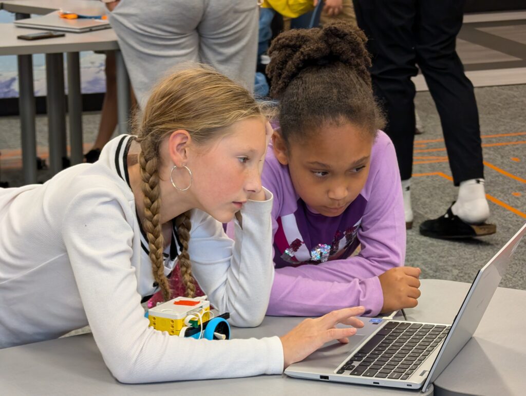 Two girls focused on a laptop screen, seated at a table, with educational materials like a small robotic device nearby.