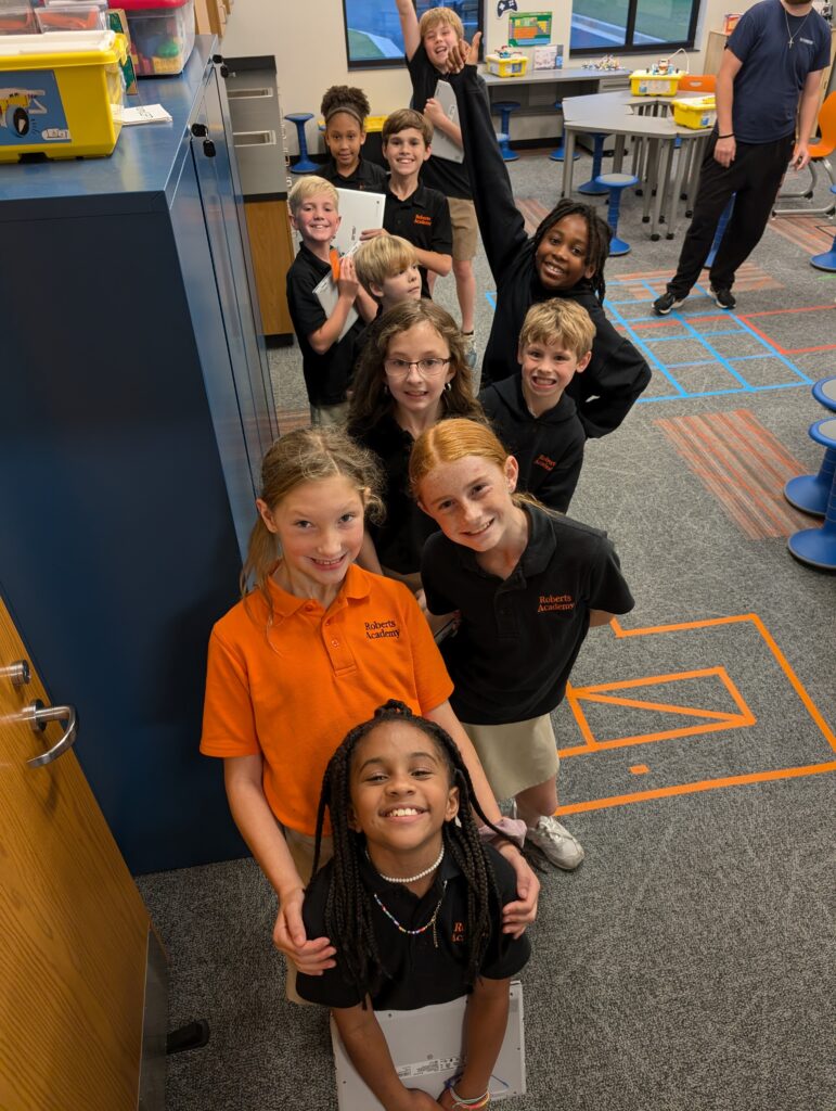 A group of smiling children in school uniforms standing in a single-file line inside a classroom.