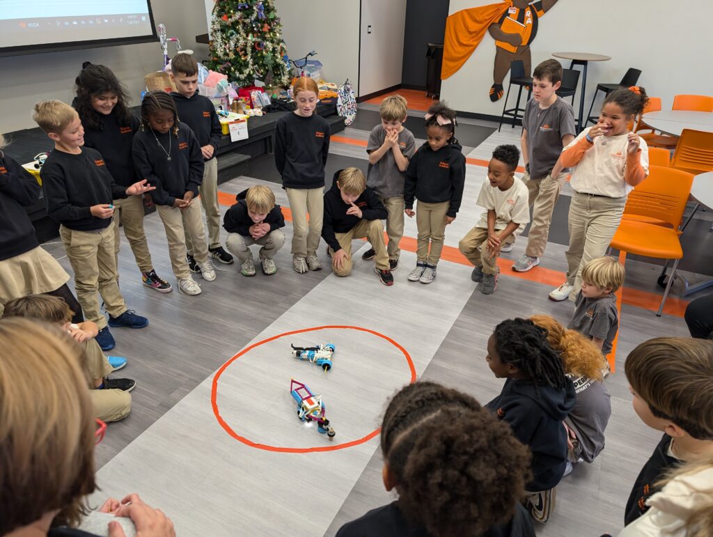 Children in a classroom excitedly watch two small robots in a taped circle on the floor.