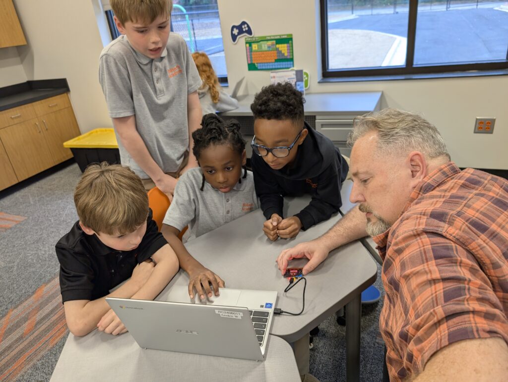 A group of children and an adult gather around a laptop on a table, engaging in a collaborative learning activity.