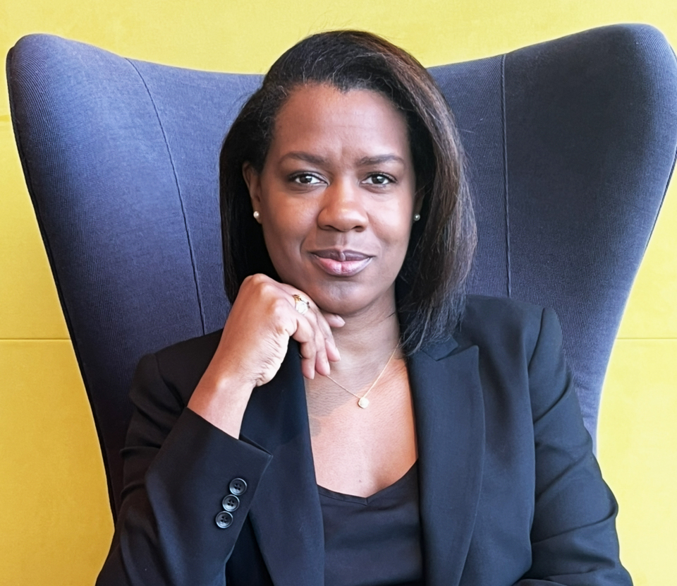 Shay Peterson in a black blazer sits in a blue chair against a yellow background.