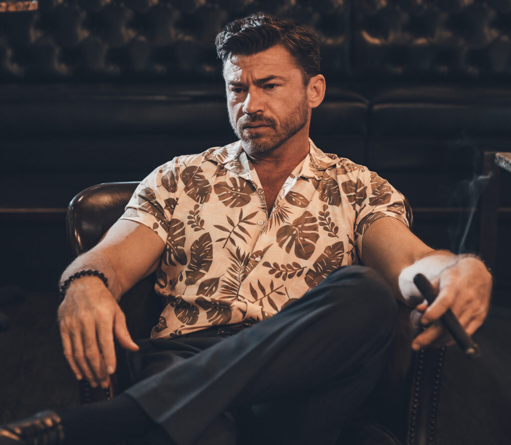 A man in a floral shirt sits in a leather chair, holding a cigar, with a serious expression in a dimly lit room.