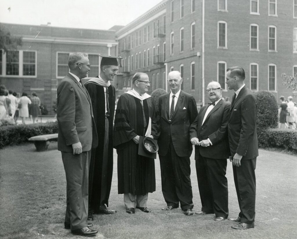 Six men, some in academic robes, stand conversing on a lawn in front of a brick building.