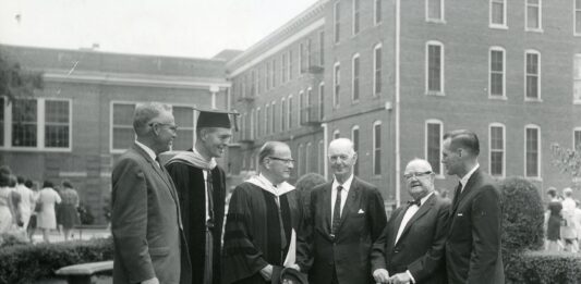 Six men, some in academic robes, stand conversing on a lawn in front of a brick building.