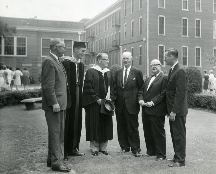 Vinson circa 1960s Six men, some in academic robes, stand conversing on a lawn in front of a brick building.