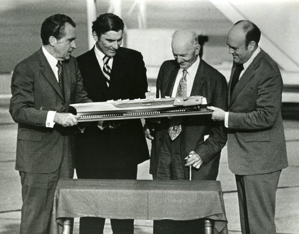 Four men in suits examining a model ship on a small table, with one man holding the ships base.