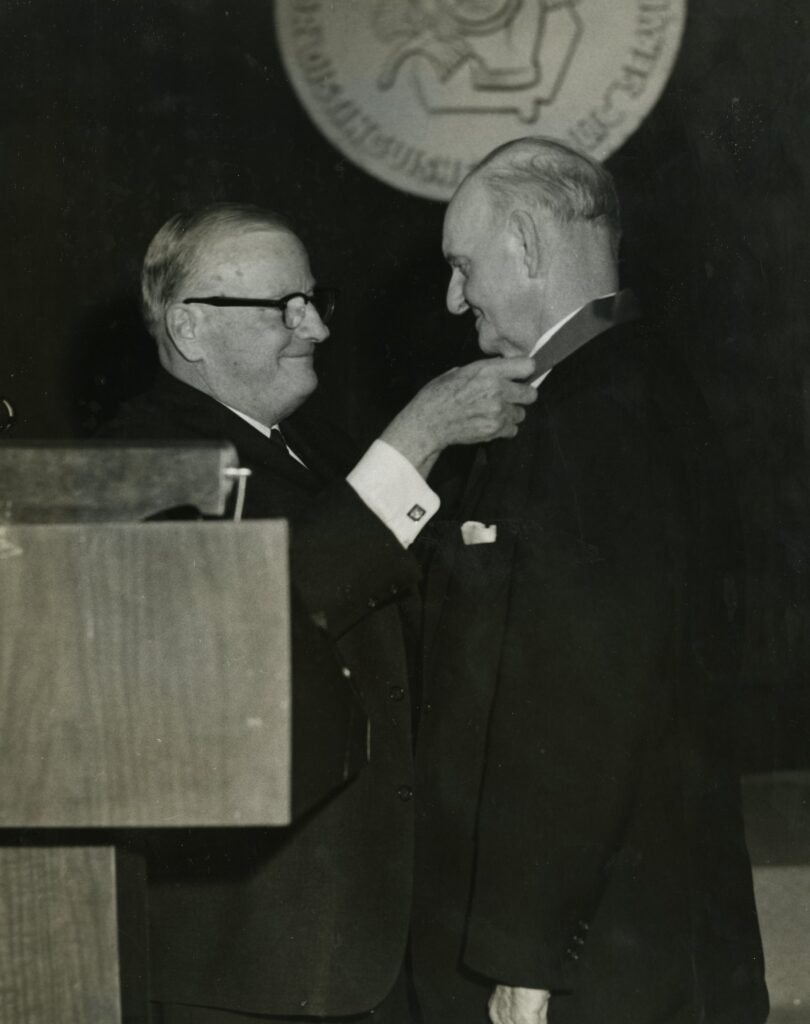 Two men in suits stand beside a podium. One man is placing a medal on the others collar. A seal is visible in the background.