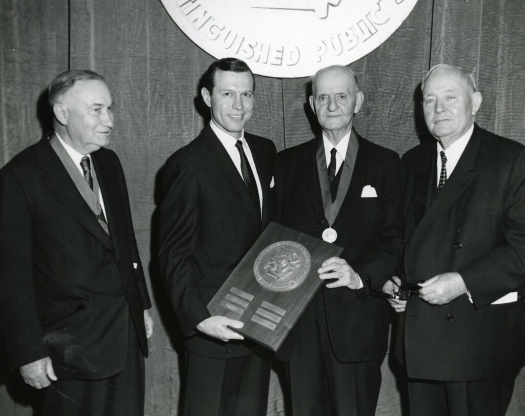 Four men in suits, one holding a plaque and another wearing a medal. They stand beneath a sign related to public service.