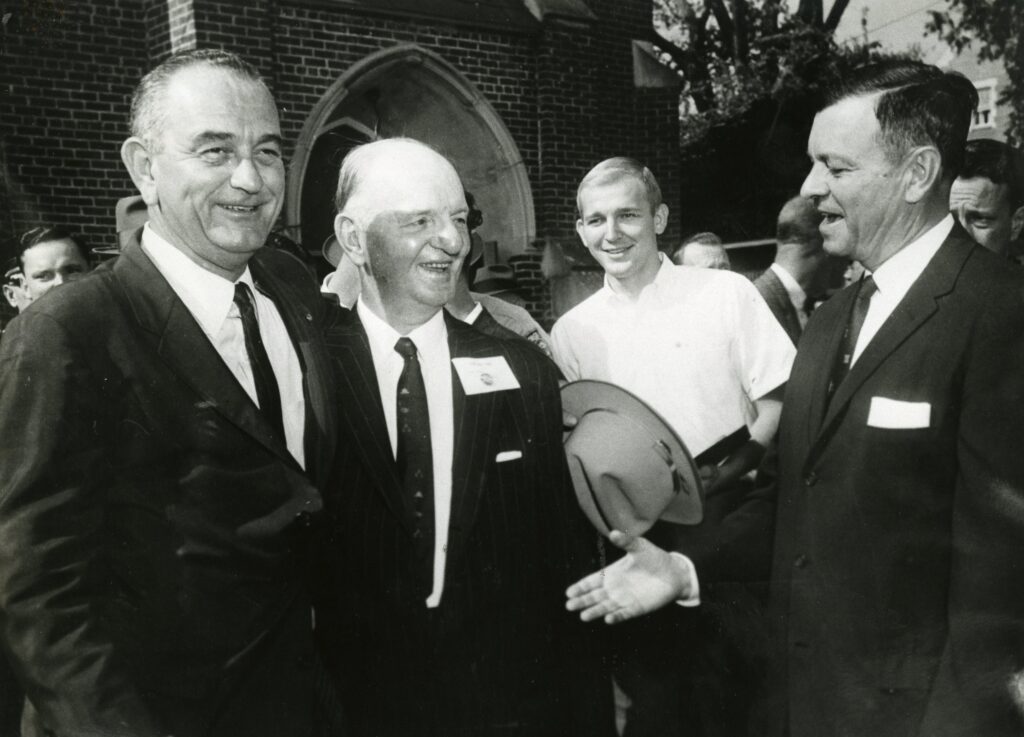 Three men in suits and a younger man in a white shirt are gathered outside a brick building, engaging in conversation and smiling.