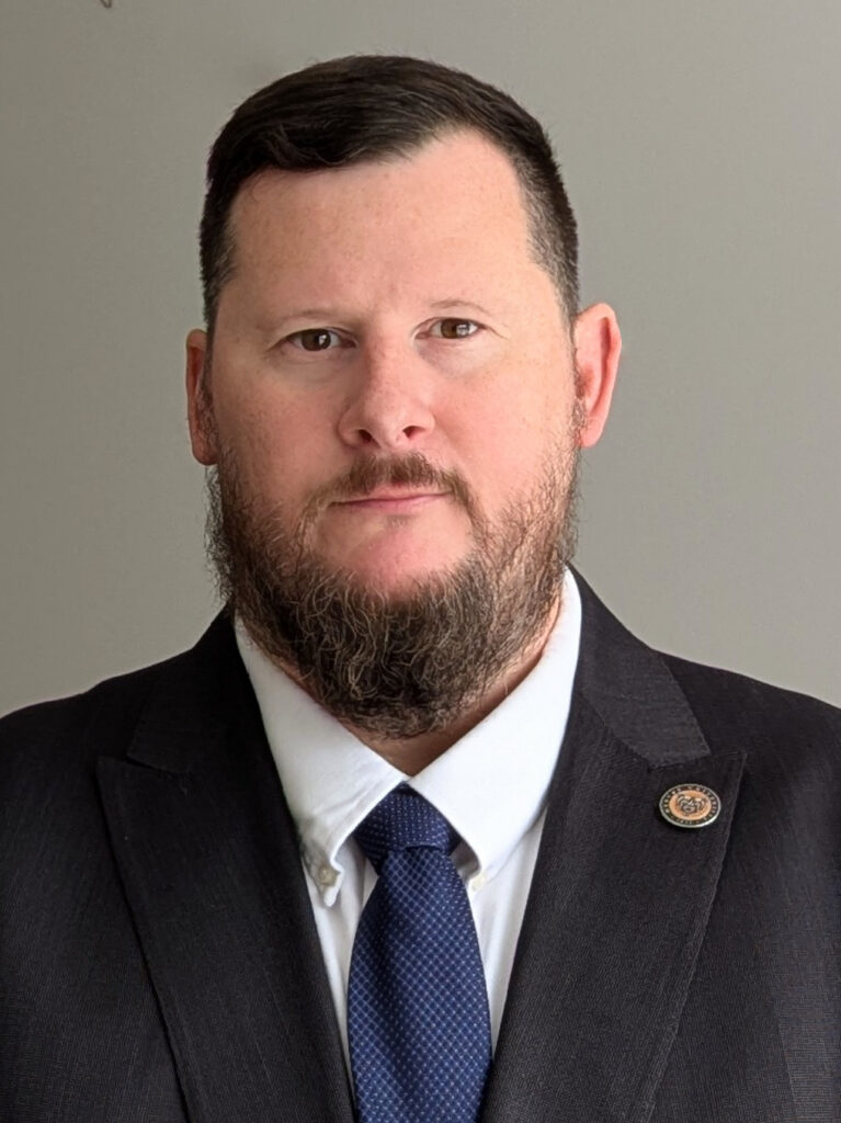 Bearded man wearing a black suit, white shirt, and navy tie, with a pin on his lapel, standing against a plain background.