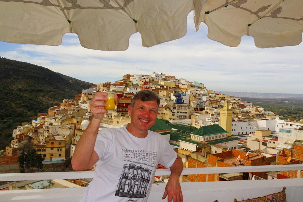Brad Lindsey holding a drink on a balcony with a view of a hillside town filled with colorful buildings under a partly cloudy sky.