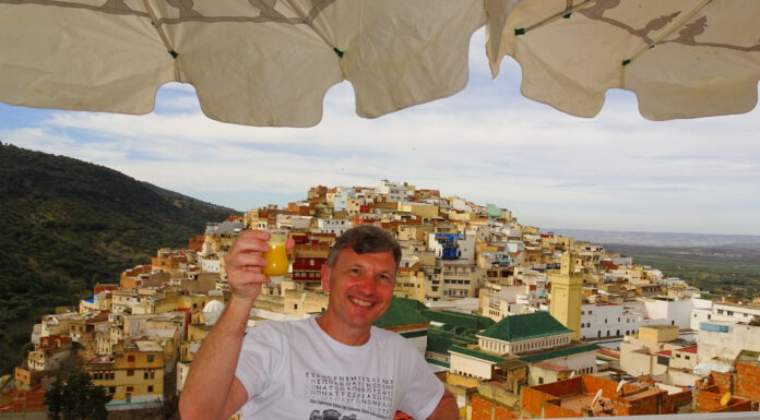 Alumnus travels to all 50 U.S. states and 7 continents Brad Lindsey holding a drink on a balcony with a view of a hillside town filled with colorful buildings under a partly cloudy sky.
