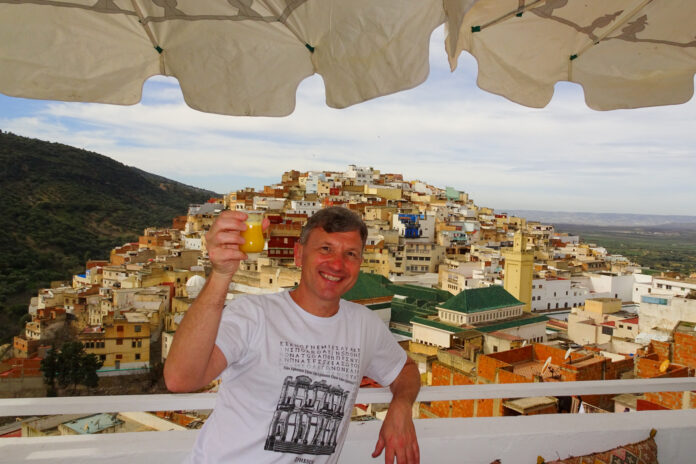 brad-lindsey-2 Brad Lindsey holding a drink on a balcony with a view of a hillside town filled with colorful buildings under a partly cloudy sky.