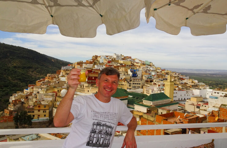 Brad Lindsey holding a drink on a balcony with a view of a hillside town filled with colorful buildings under a partly cloudy sky.