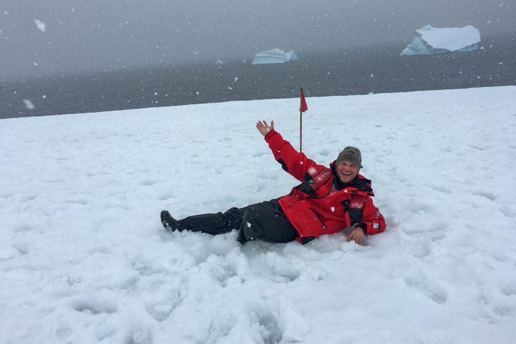Brad Lindsey lies on snowy ground near the ocean with icebergs in the background.