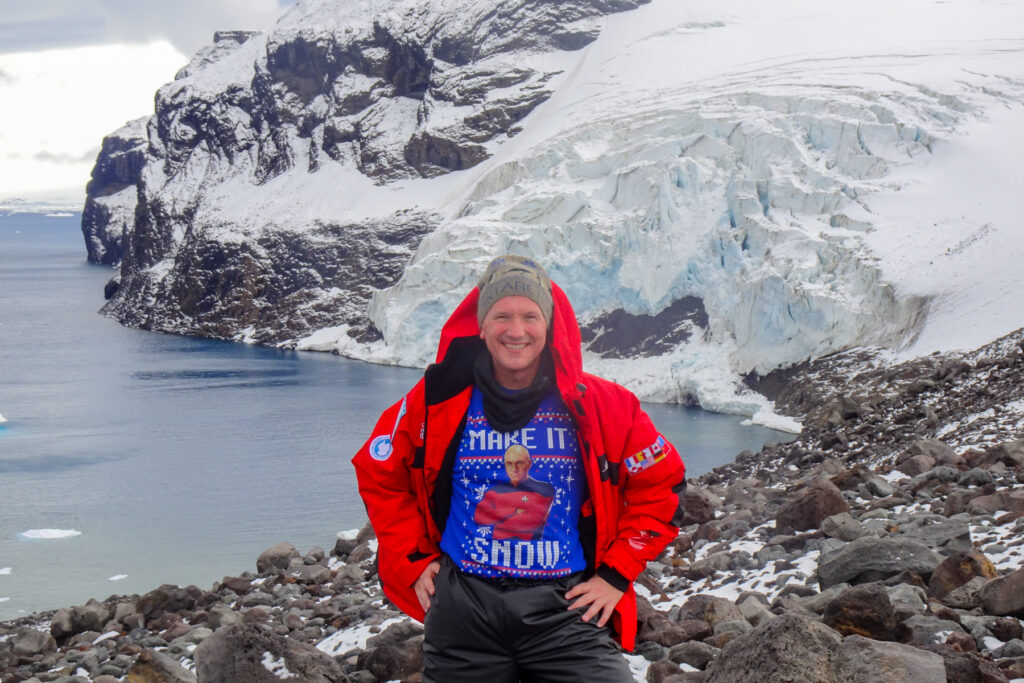 Brad Lindsey stands on rocky terrain near a snowy cliff and icy water, with glaciers in the background.