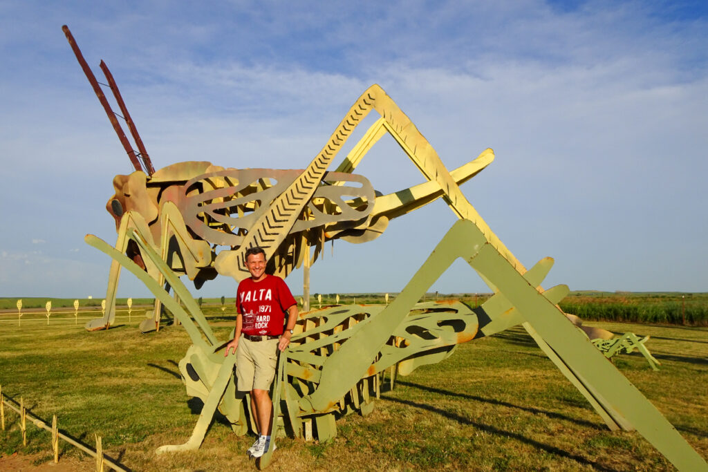 Brad Lindsey standing next to a large, metal grasshopper sculpture on a grassy field under a blue sky.