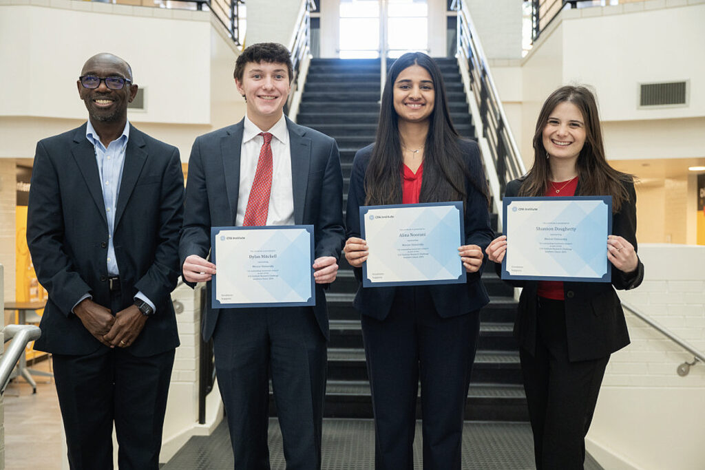 Four people stand indoors; three hold certificates. They are dressed in business attire and stand in front of a staircase.