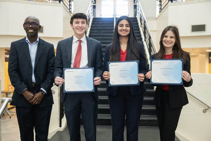 Four people stand indoors; three hold certificates. They are dressed in business attire and stand in front of a staircase.