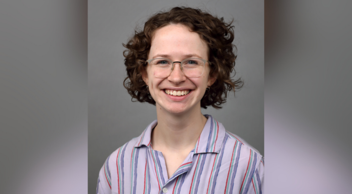 Junior hones research skills, focus at Mercer Person with curly hair and glasses smiling, wearing a striped shirt against a plain background.