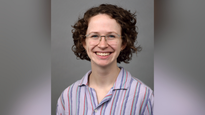 Person with curly hair and glasses smiling, wearing a striped shirt against a plain background.
