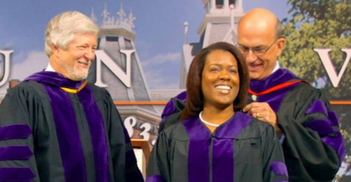 Three people in purple academic robes at a graduation ceremony; a woman is being honored while two men stand beside her, smiling.