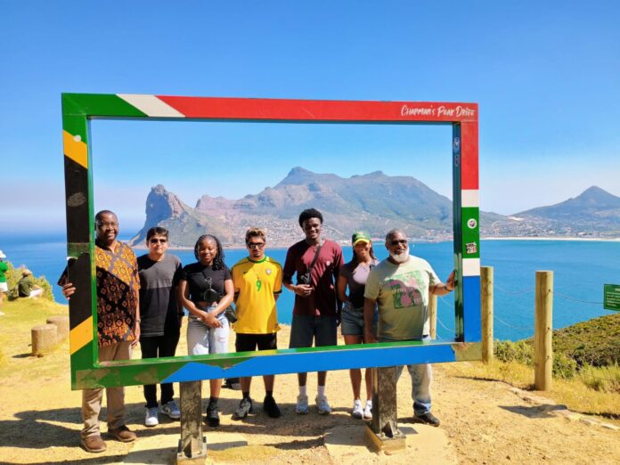 Group of seven people standing in a colorful frame with a coastal landscape backdrop and mountains visible in the distance.