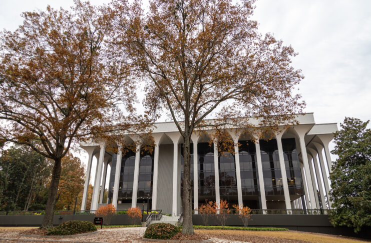 Modern building with tall white columns and large glass windows, framed by autumn trees with orange and brown leaves.