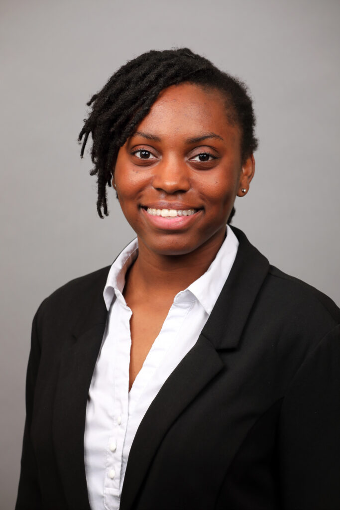 A person in a black blazer and white shirt smiles at the camera against a plain gray background.