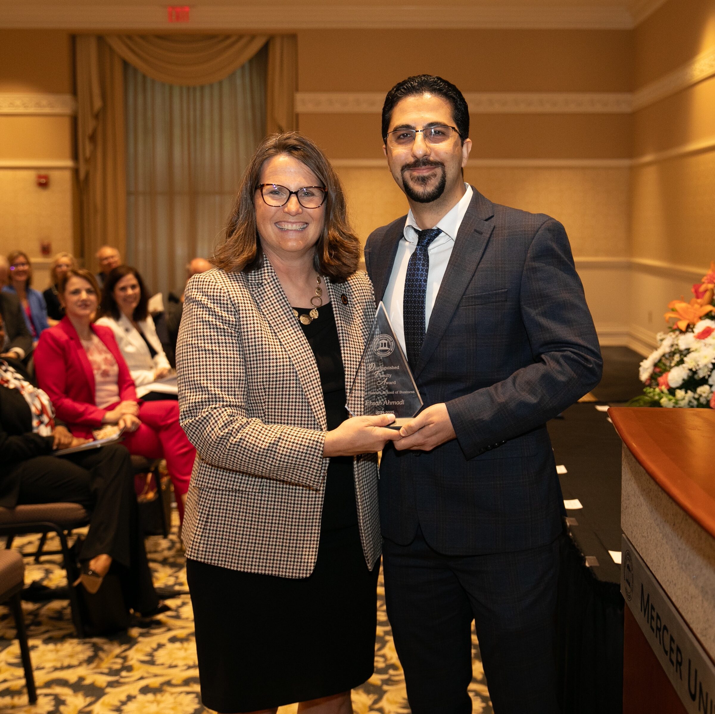 Two people stand smiling with an award in a formal setting, audience seated in the background.