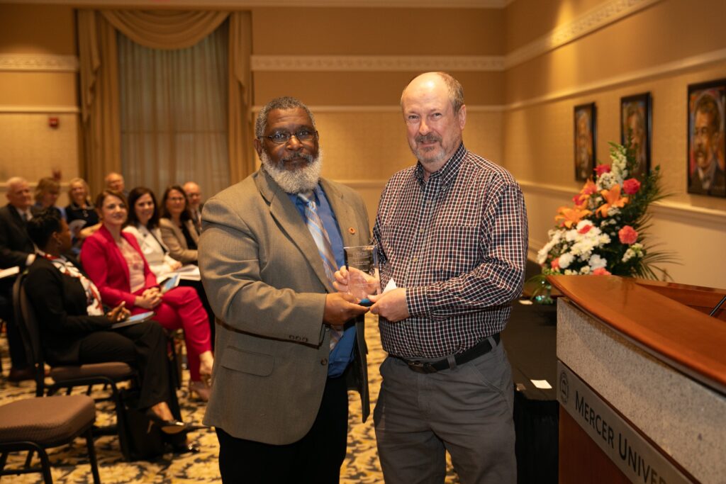 Two men shaking hands during an award ceremony in a conference room, with an audience seated in the background.