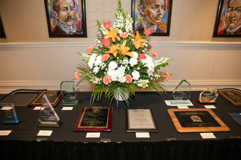 A display of various plaques and awards on a black tablecloth, with a floral arrangement in the center against a background of paintings.