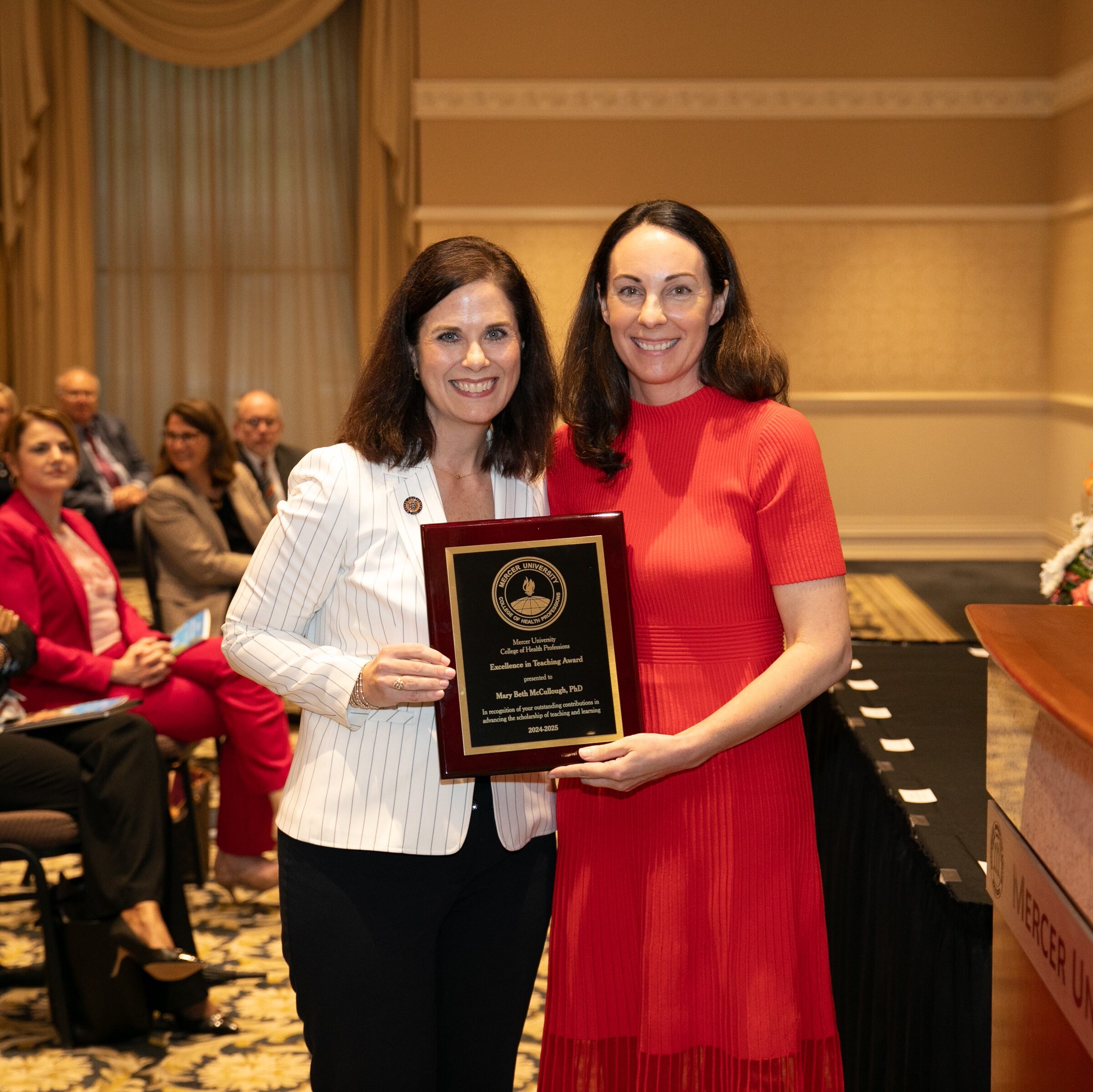 Two women stand in front of an audience. One holds an award plaque. They are both smiling. Flowers and podium are visible.