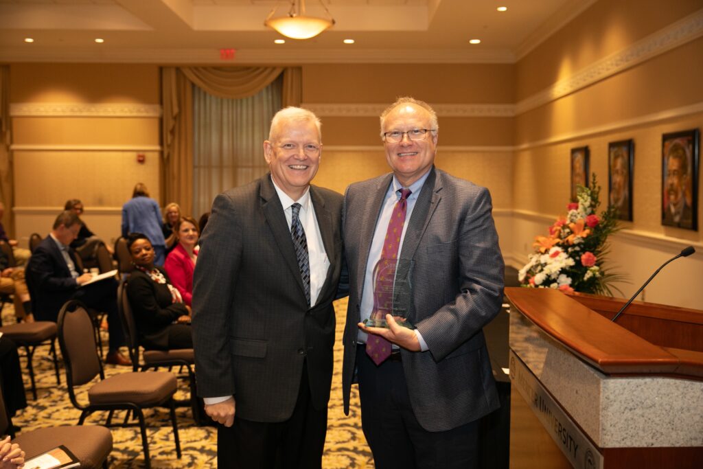 Two men in formal attire pose for a photo in a conference room. One holds an award. People sit in the background.
