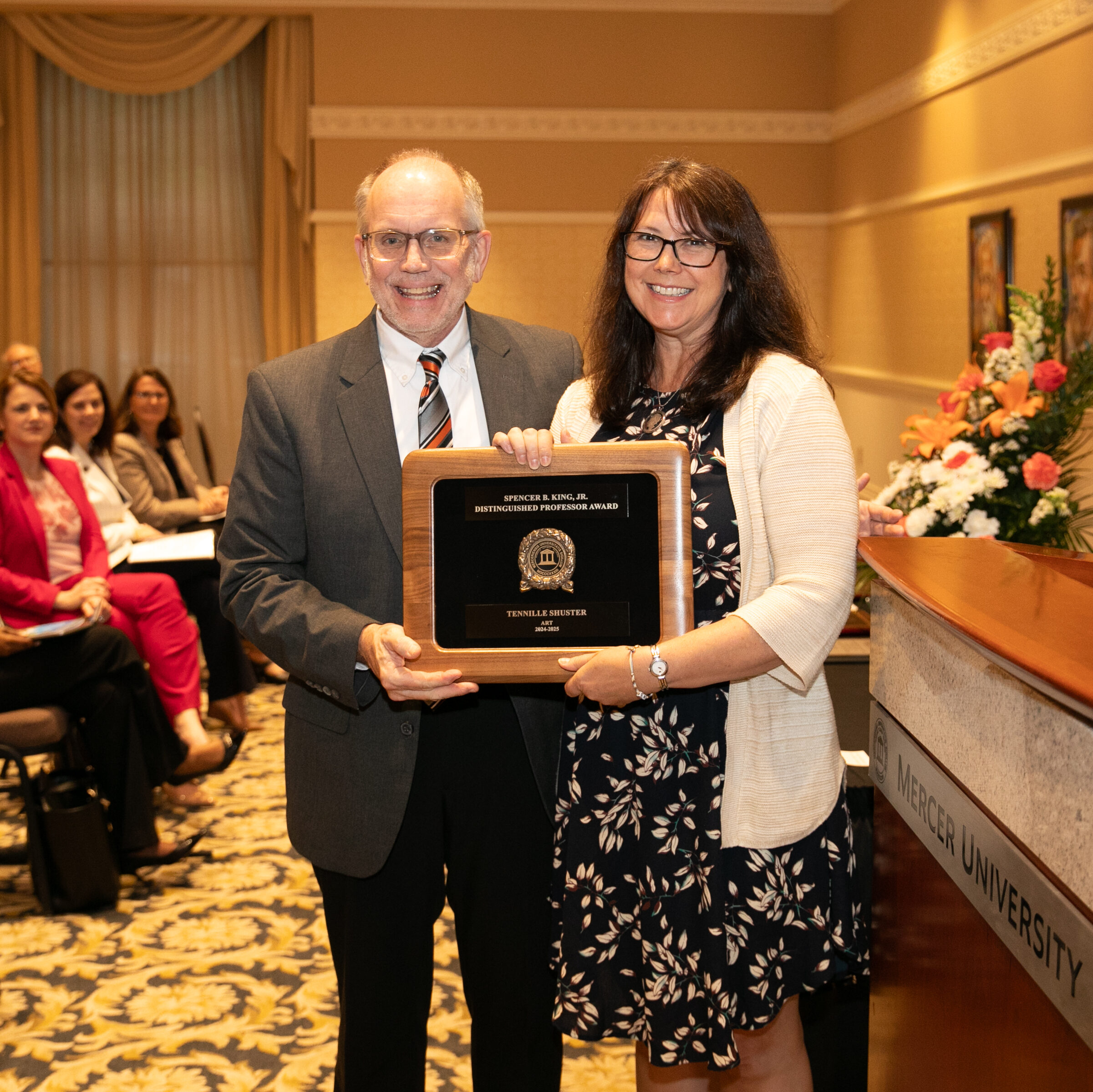 Two people holding an award plaque in a conference room, with an audience seated behind them and a lectern nearby.