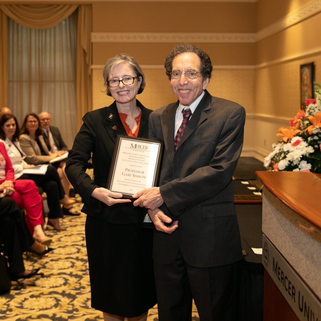 Two people stand smiling, holding an award plaque, in a formal setting with seated audience and floral arrangement.