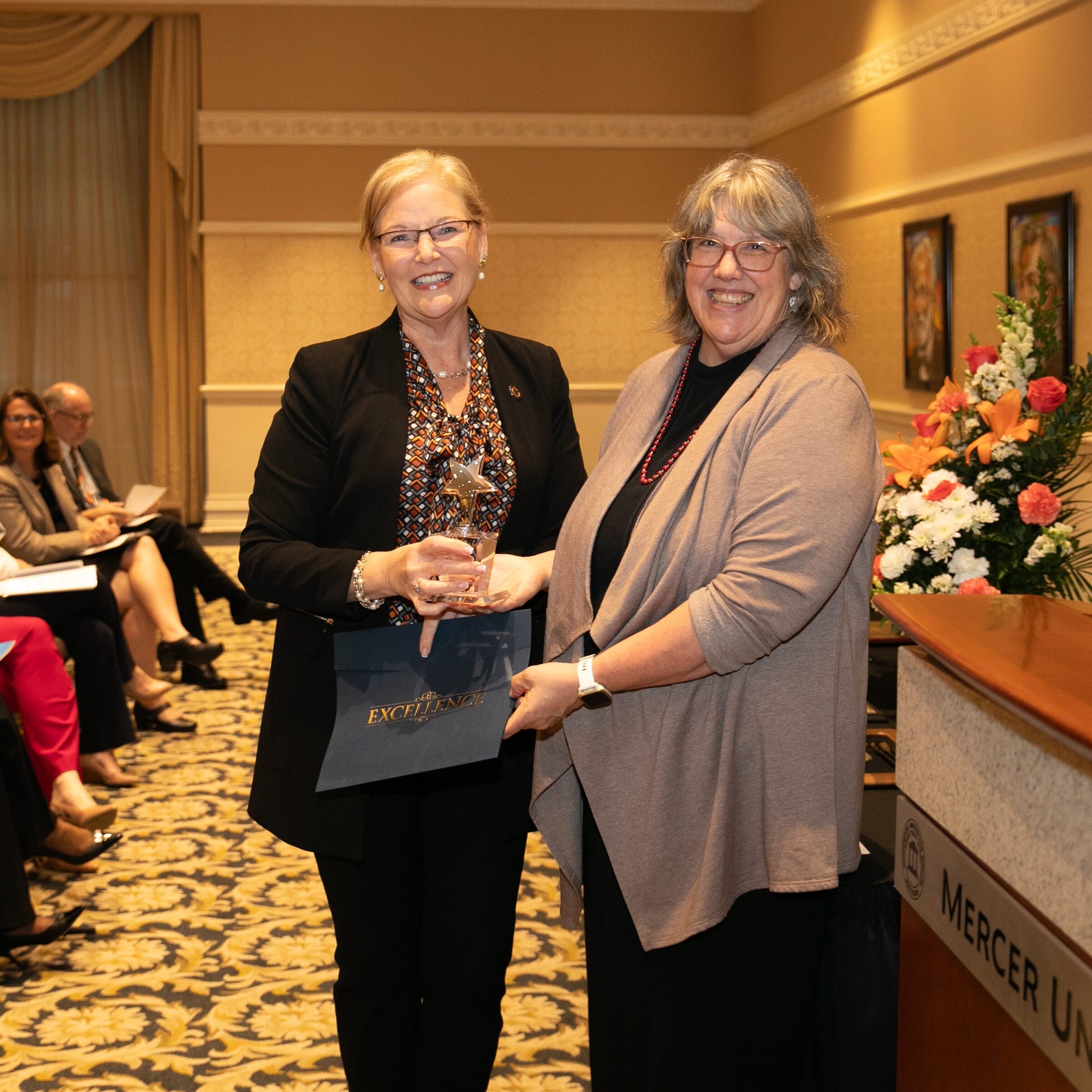 Two people smiling and holding an award certificate in a conference room, with an audience seated in the background.