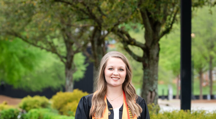 Graduate Spotlight: Allison Slaughter Allison Slaughter in graduation gown and stole stands holding cap on a sidewalk lined with greenery and trees.