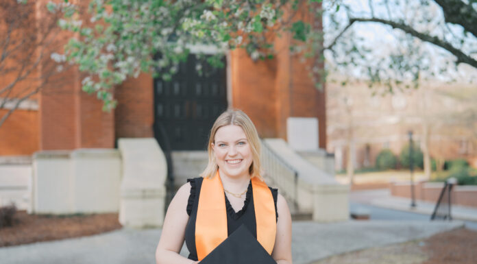 Graduate Spotlight: Anna Hale Anna Hale in a black graduation outfit holds a cap outdoors, standing in front of a brick building and trees.