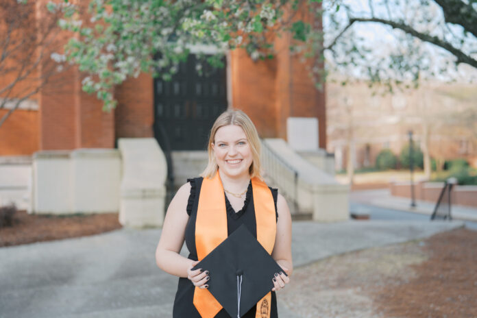 Anna Hale in a black graduation outfit holds a cap outdoors, standing in front of a brick building and trees.