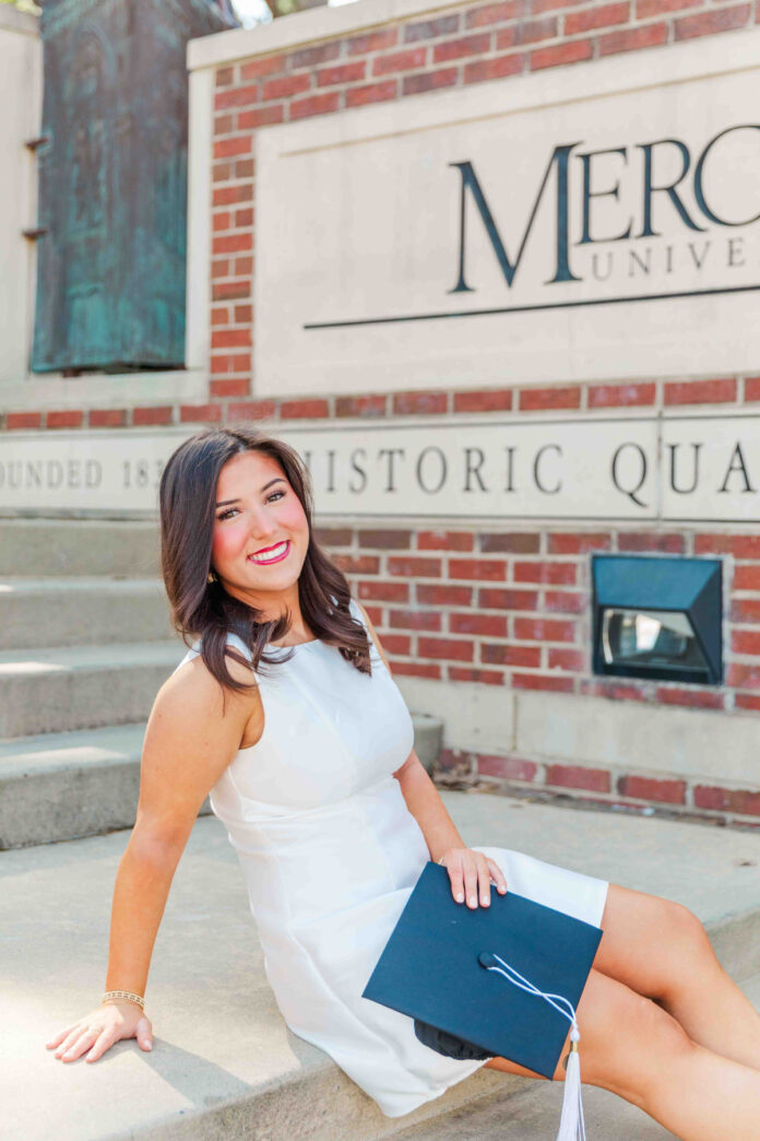 Cameron White, in a white dress, sits on steps holding a cap, with a Mercer University sign in the background.