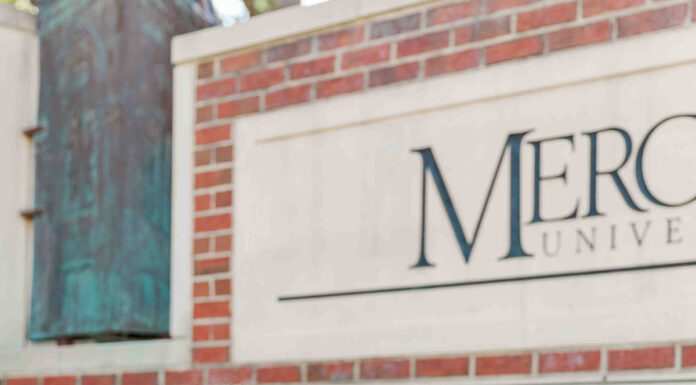 Graduate Spotlight: Cameron White Cameron White, in a white dress, sits on steps holding a cap, with a Mercer University sign in the background.
