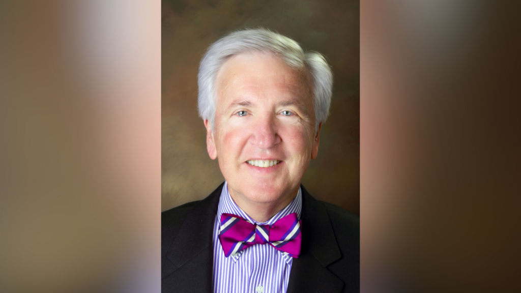 A person with gray hair wearing a suit, colorful striped shirt, and bright purple bow tie, smiling at the camera.
