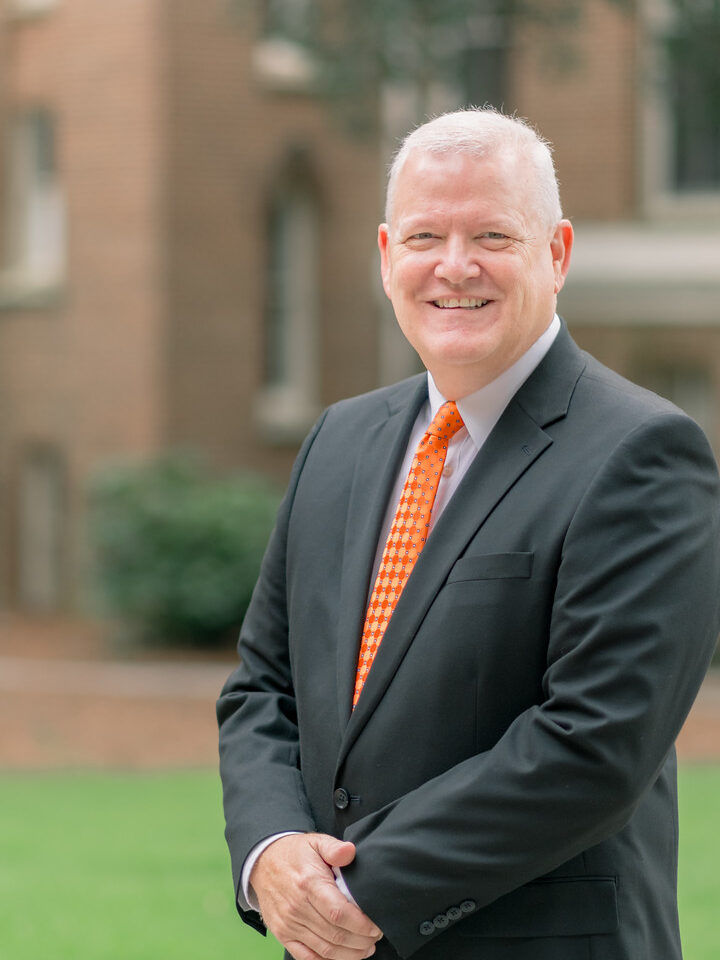 A man in a black suit and orange tie stands outdoors, smiling, with a blurred building in the background.