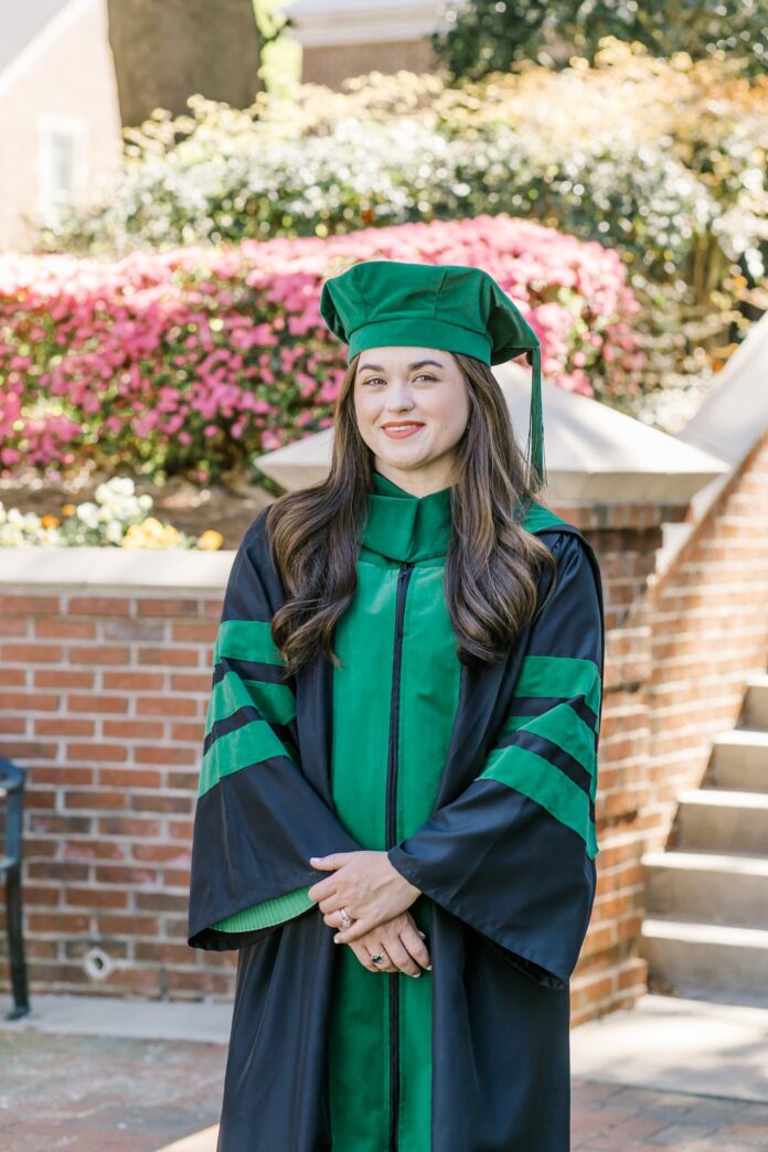 Elizabeth Pitman, in green cap and gown, stands outdoors in front of blooming bushes and a brick wall.